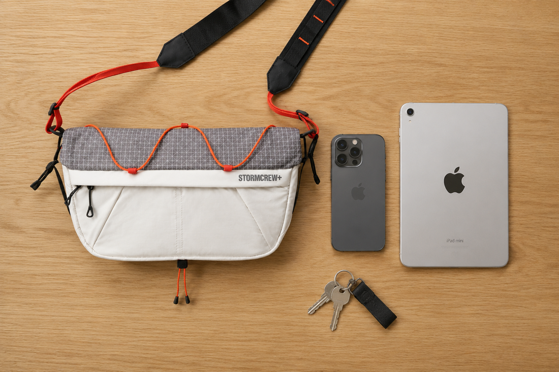 Minimalist flat lay comparing a white Stormcrew sling bag size to an iPad Mini, iPhone, and keys on an oak desk.